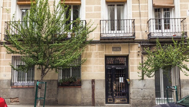 A building with a traditional facade, featuring a central entrance with a black gate, balconies with wrought iron railings, and a tree in front of the entrance.