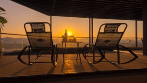 Deux fauteuils à bascule placés face à un point de vue offrant un magnifique coucher de soleil, sur un balcon agrémenté d’une table et de verres à vin.