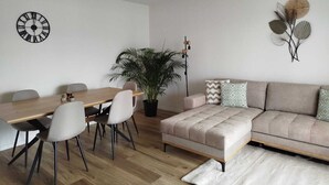 Een moderne woonkamer met een houten eettafel, witte stoelen, een beige bank, een grote potplant en decoratieve muurkunst.