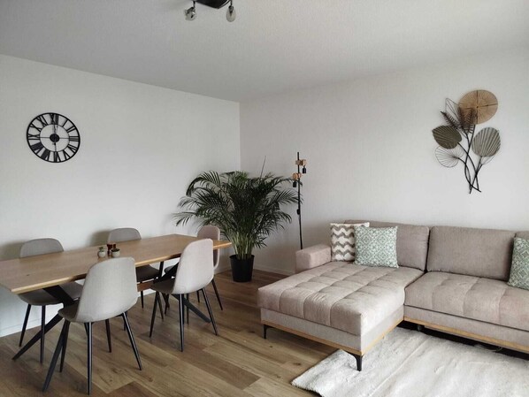 Un salon moderne avec une table à manger en bois, des chaises blanches, un canapé beige, une grande plante en pot et des œuvres d’art décoratives accrochées au mur.