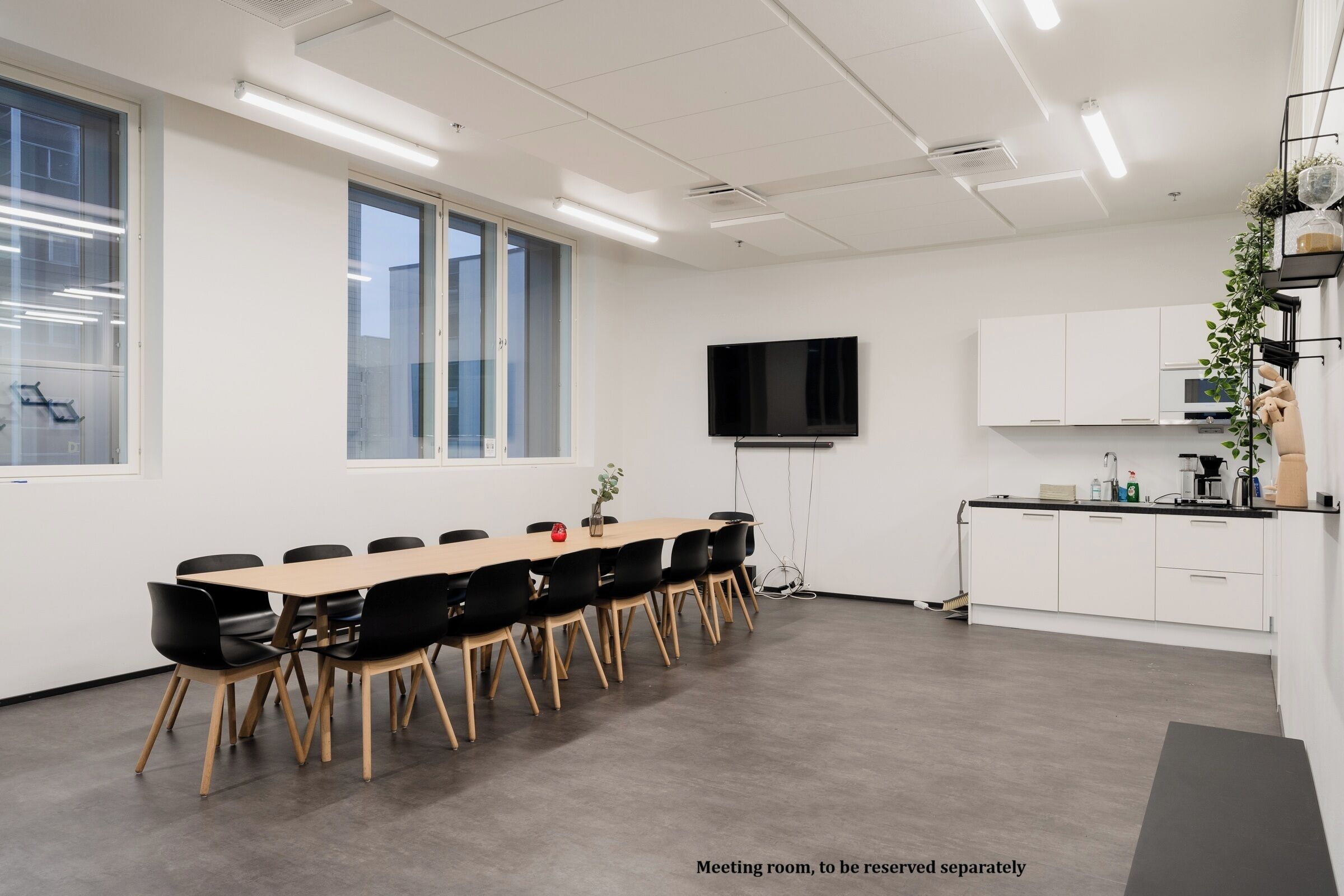 A meeting room with a long table, black chairs, a flat-screen TV, and a kitchenette.