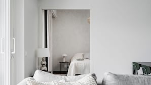 Un dormitorio moderno y minimalista con un sofá gris, una cama blanca y una mesita de noche con lámpara.