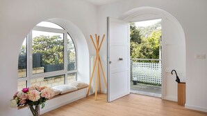 Una stanza con un'ampia finestra ad arco, una panca di legno, un vaso di fiori e una porta che dà sull'esterno.