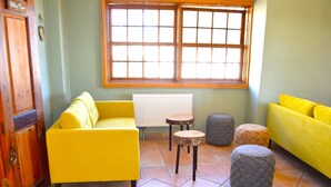 Una stanza con un divano giallo, un armadietto in legno, un termosifone, una finestra con persiane in legno e due sgabelli rotondi.