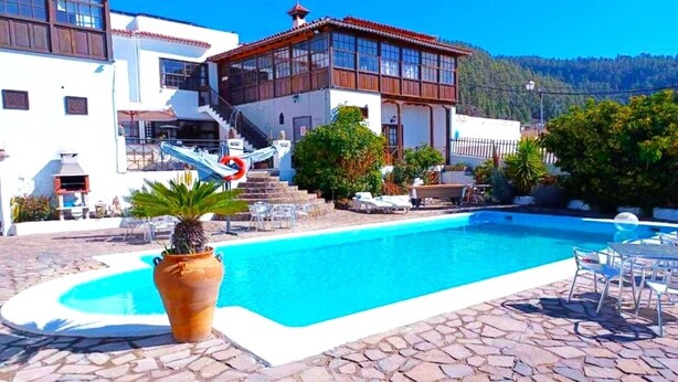 Una piscina con bordo in pietra, circondata da mobili da esterno e un edificio con tetto in tegole rosse.