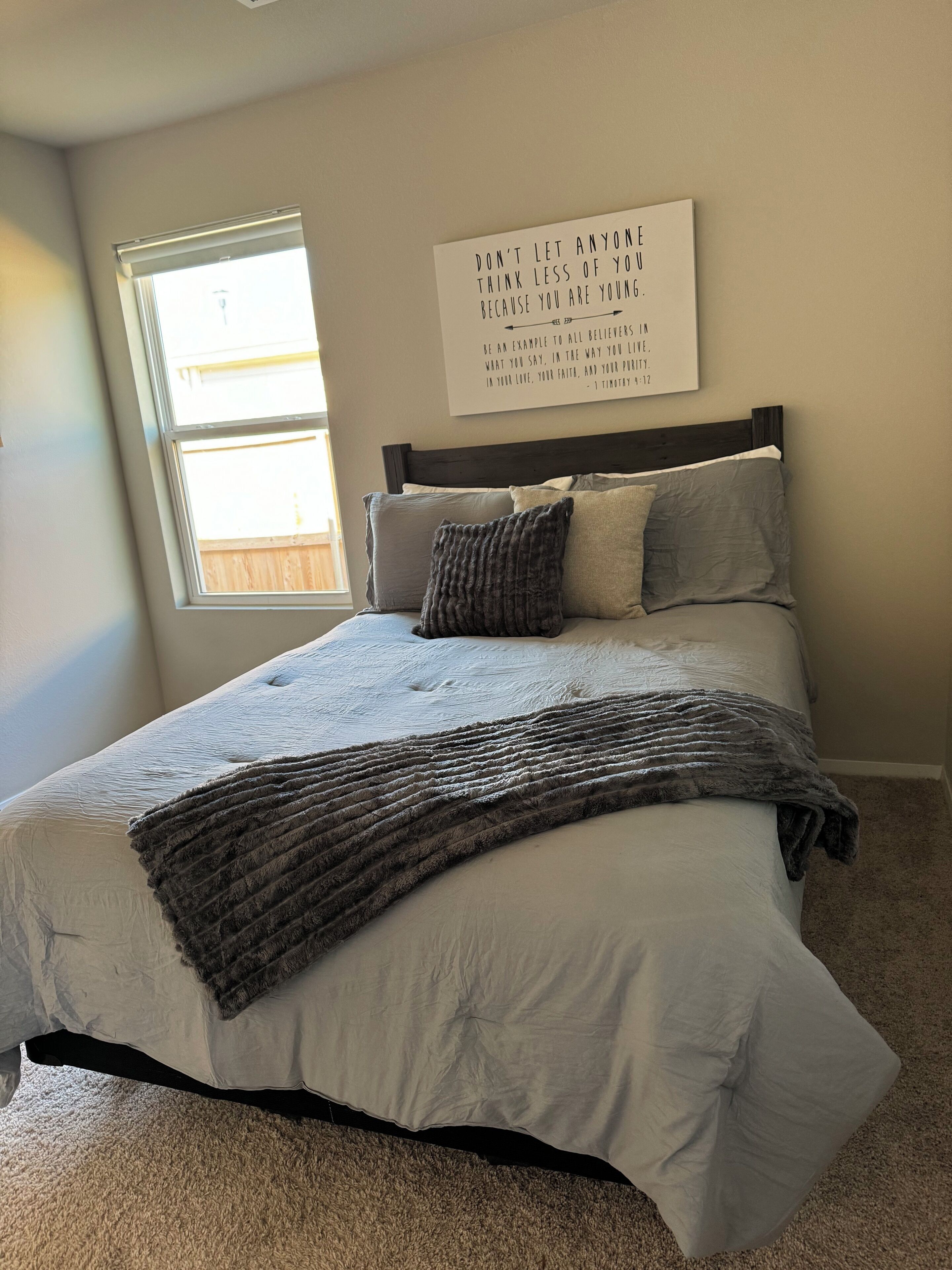 A bedroom with a bed, pillows, a blanket, a window, and a motivational quote on the wall.