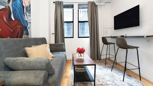 Ein modernes Wohnzimmer mit einem grauen Sofa, einem Couchtisch mit einer Vase roter Blumen und zwei Barhocker. Ein großes Fenster mit Vorhängen, ein Flachbildfernseher an der Wand und ein Gemälde sind ebenfalls zu sehen.