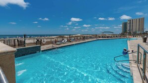 A swimming pool with clear blue water, surrounded by lounge chairs and umbrellas, with a beach and hotel buildings in the background.