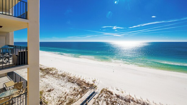 A beachfront hotel with a balcony overlooking the ocean, a sandy beach, and clear skies.
