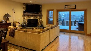 A cozy living room with a stone fireplace, a large flat-screen TV, a comfortable sofa, and a view of a snowy deck through glass doors. - Spacious 4-bedroom Ski- in/ Ski-out condo in enchanting Sunday River w/ Hot Tub  (Newry)