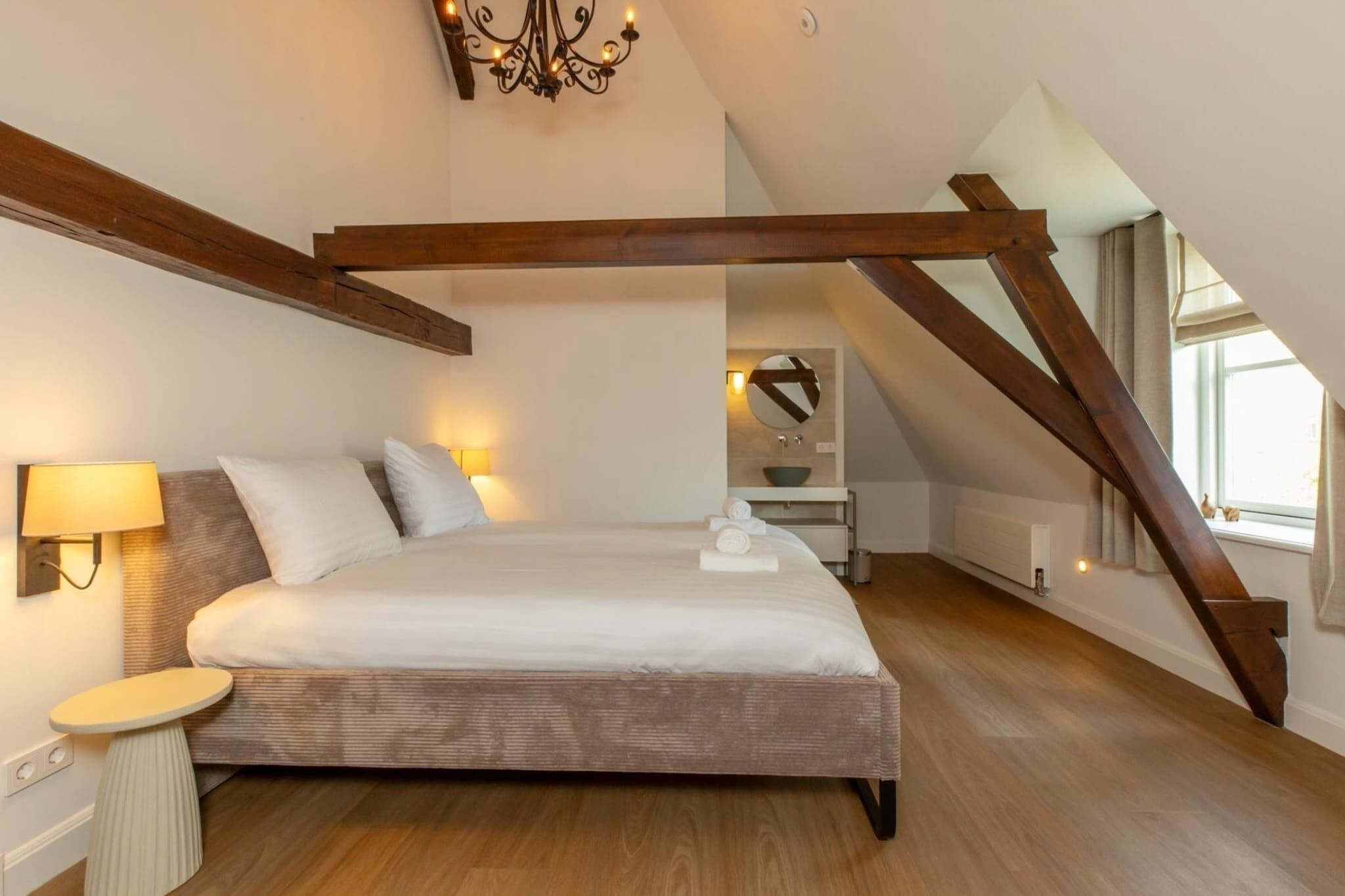A bedroom with a large bed, a bedside table with a lamp, a chandelier, and a view of a bathroom through an open door.