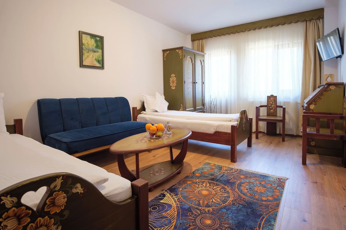 Ein Hotelzimmer mit blauem Sofa, einem Bett mit weißer Bettwäsche, einem Holzschrank, einem kleinen Tisch mit Obst und einem gemusterten Teppich.