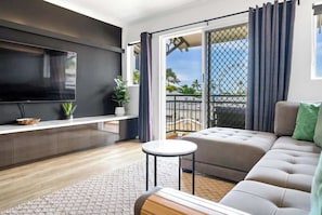 A modern living room with a flat-screen TV, a sofa, a small round table, and a balcony with curtains.