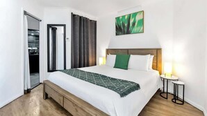 A hotel room with a bed, a bench, a small table, a painting, and a door leading to a kitchen.