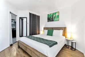 Una camera d'hotel con un letto, una panca, un tavolino, un quadro e una porta che dà su una cucina.