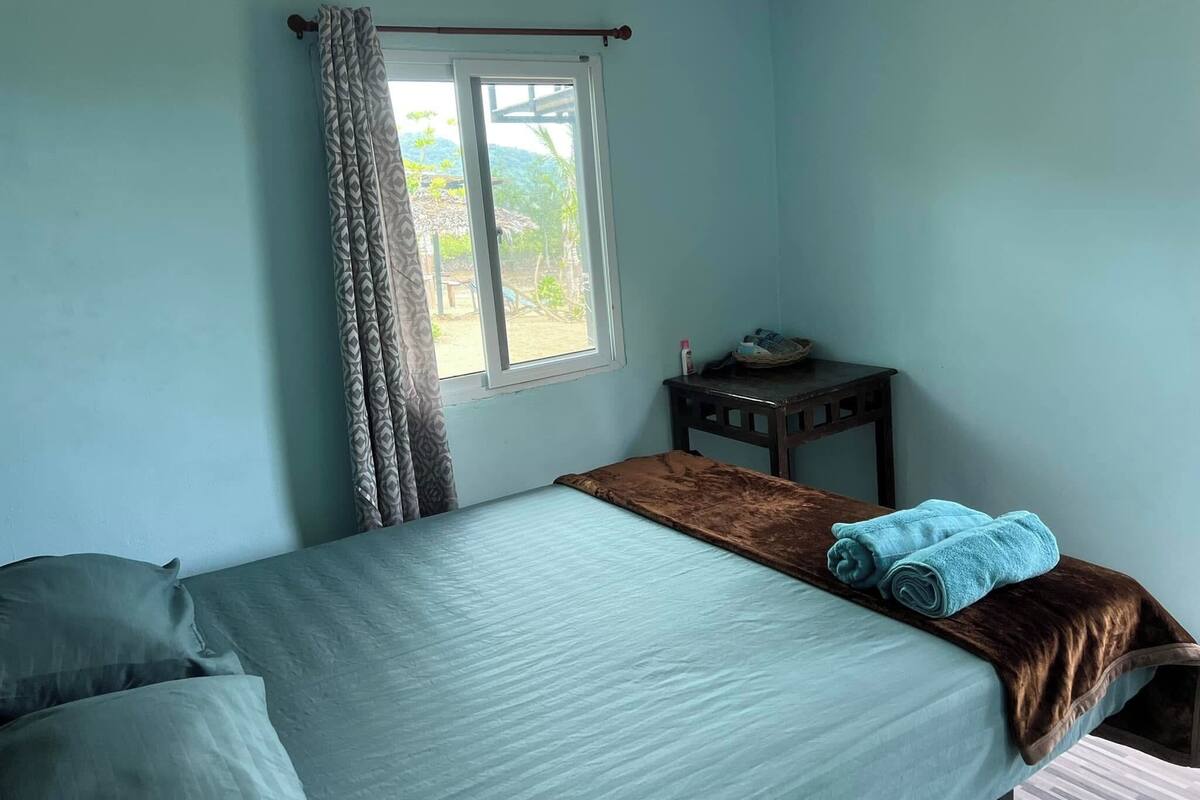 A bedroom with a bed, a window with curtains, a small table with a shoe and a bottle, and a view of greenery outside.