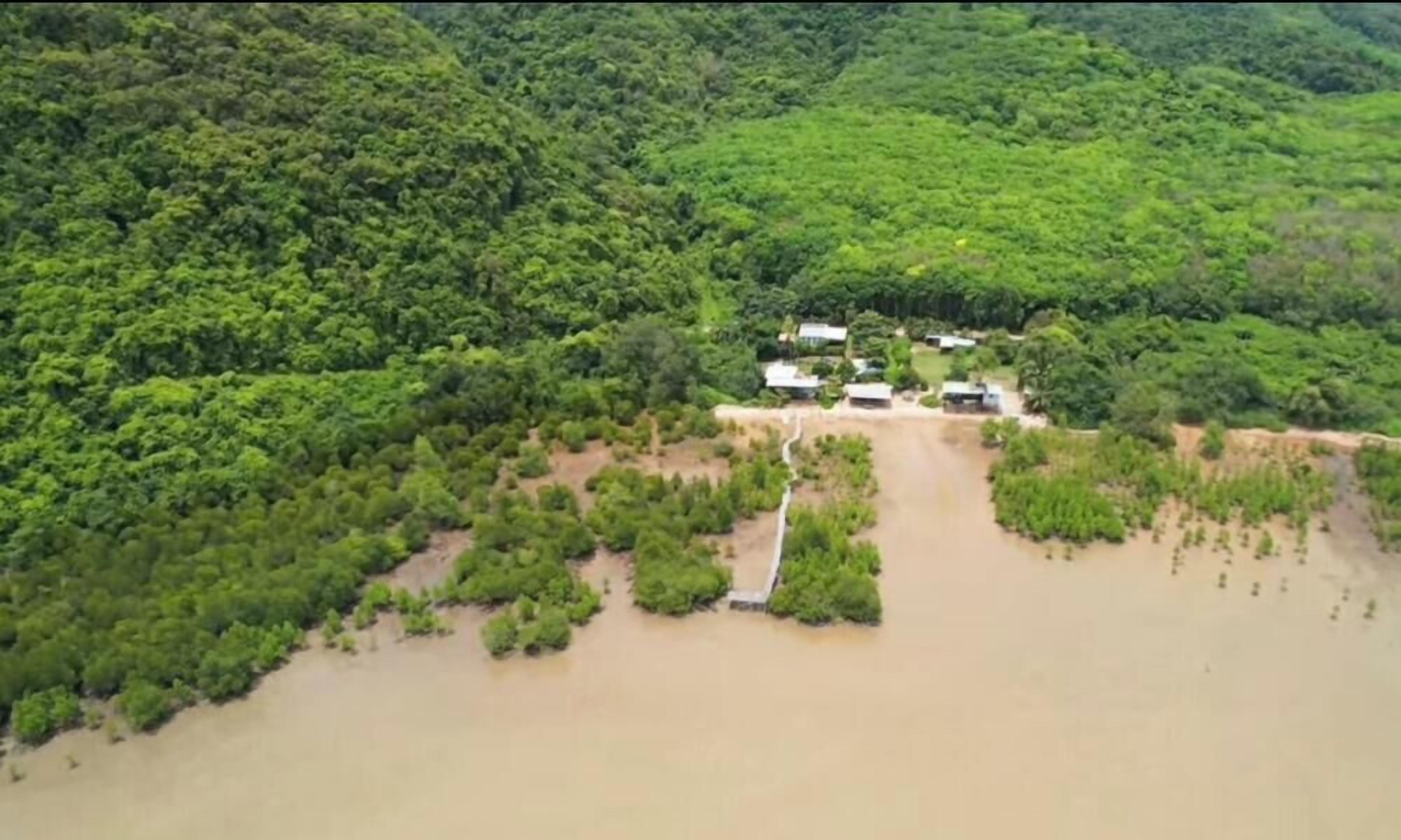 An aerial view of a property surrounded by dense forest, with a water body in the foreground and a few buildings visible.
