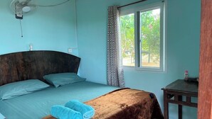 A bedroom with a bed, a bedside table, a fan, a window with curtains, and a view of greenery outside. - Baan Plai Laem Resort (Ko Phaluai)