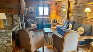 Interior rústico de una cabaña con pared de piedra, techo de madera y una zona de estar acogedora con un sofá y dos sillones.