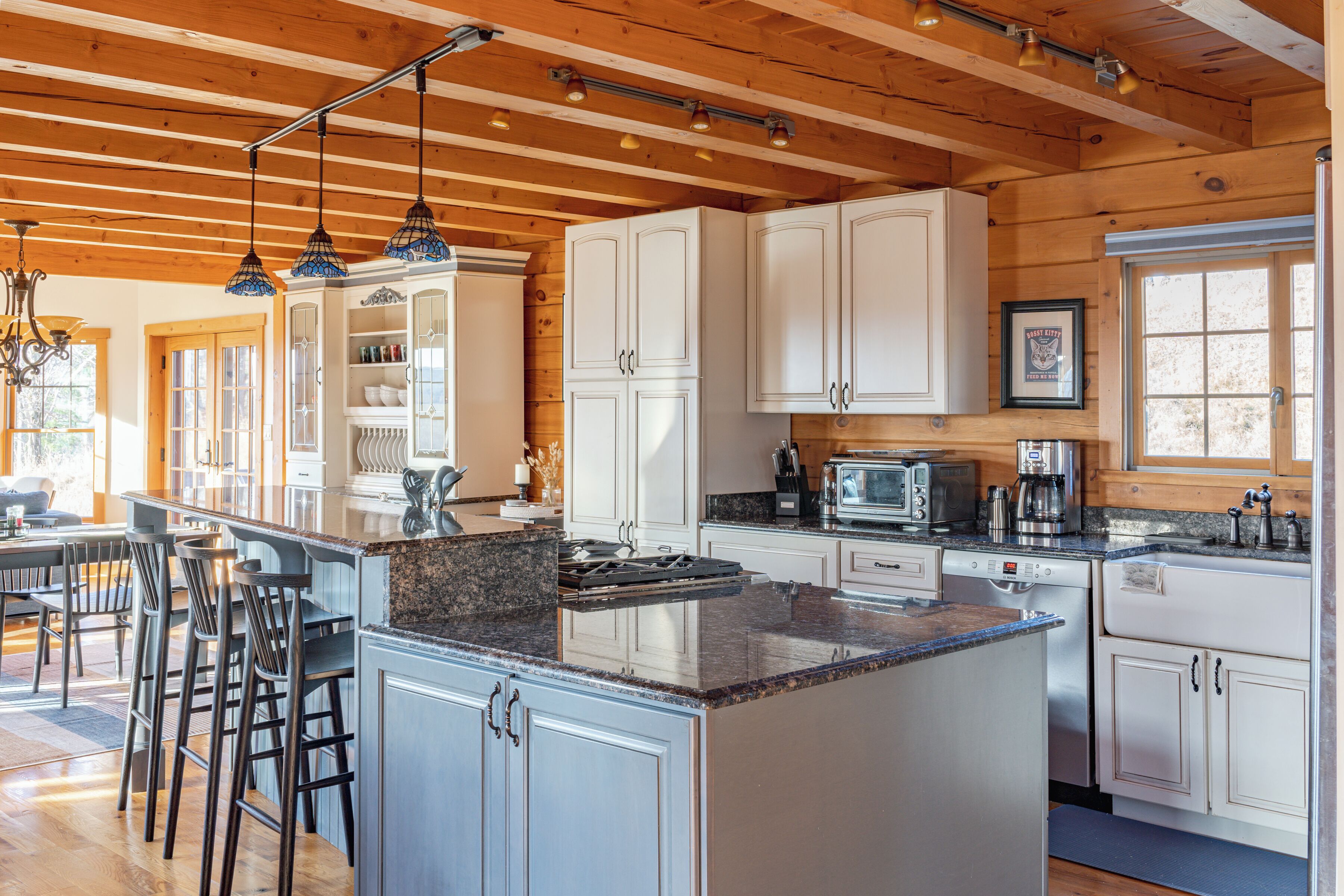 A spacious kitchen with wooden beams, white cabinets, a central island, bar stools, and a dining area with a large window.