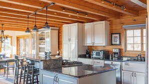 A spacious kitchen with wooden beams, white cabinets, a central island, bar stools, and a dining area with a large window.