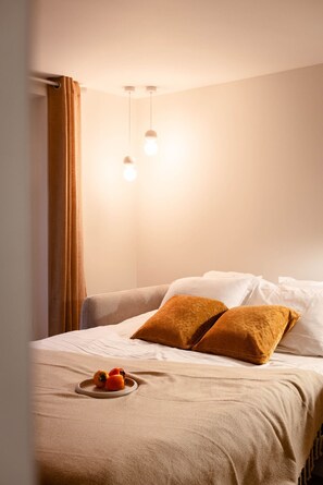 Un lit bien fait, recouvert d’une couverture beige, avec deux coussins à motifs et une assiette de fruits posée sur le lit.
