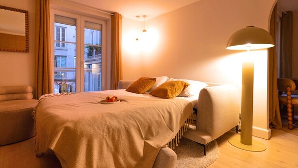 Quarto de hotel com cama, luminária de cabeceira, cadeira, mesinha com frutas e vista para prédios pela janela.