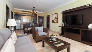 Ein Wohnzimmer mit Sofa, Sessel, Couchtisch, Fernseher und einer Küche im Hintergrund.
