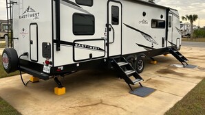 Een witte camper met 'EAST WEST Alta' branding, een ladder en een hellingbaan voor toegankelijkheid.