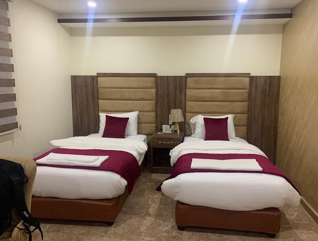 A hotel room with two beds, each with a headboard, a nightstand, and a lamp.