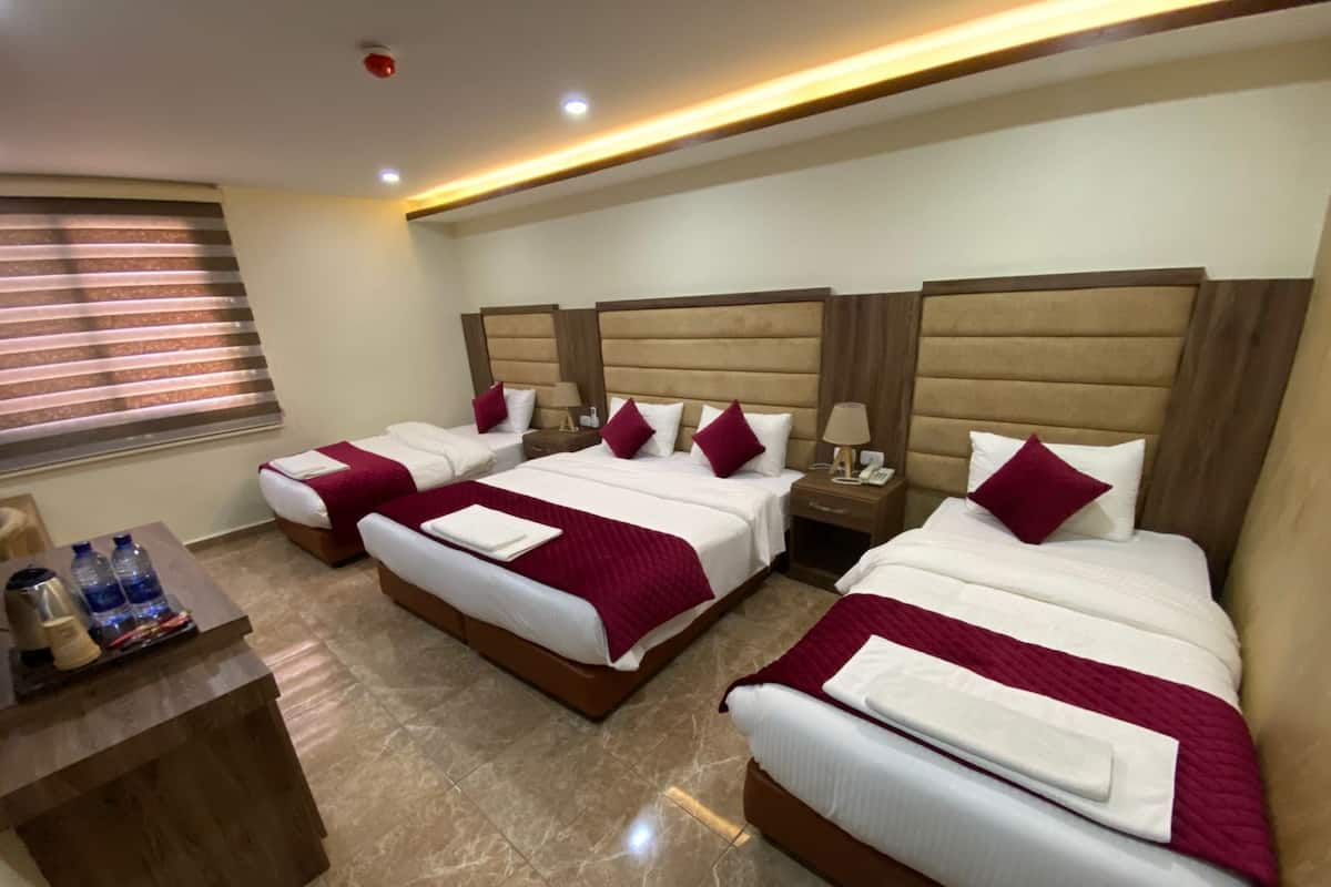 A hotel room with three beds, each with a wooden headboard, white bedding, and red throw pillows. There is a wooden nightstand with a lamp and a phone between the beds. A small table with water bottles and a coffee maker is visible.