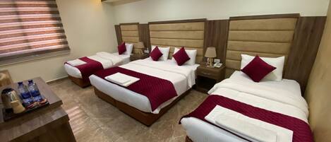 A hotel room with three beds, each with a wooden headboard, white bedding, and red throw pillows. There is a wooden nightstand with a lamp and a phone between the beds. A small table with water bottles and a coffee maker is visible.