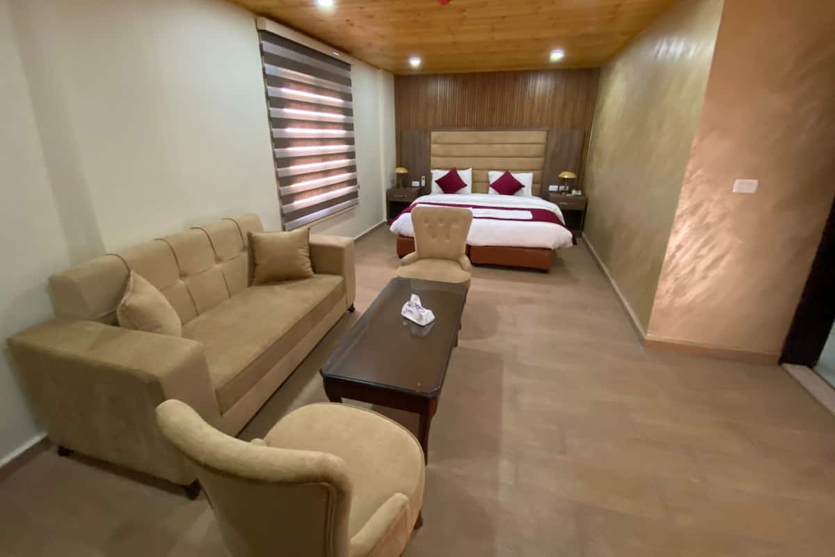 A hotel room with a wooden ceiling, a large bed with red pillows, a beige sofa, a small armchair, and a nightstand with a lamp.