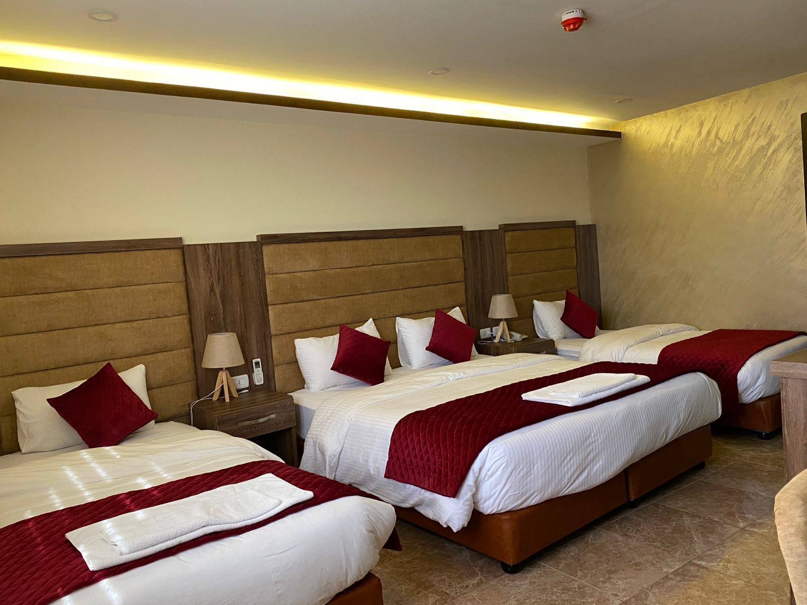 A hotel room with four beds, each with white bedding and red accents, wooden headboards, and bedside tables with lamps.