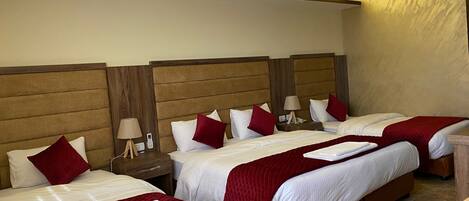 A hotel room with four beds, each with white bedding and red accents, wooden headboards, and bedside tables with lamps.