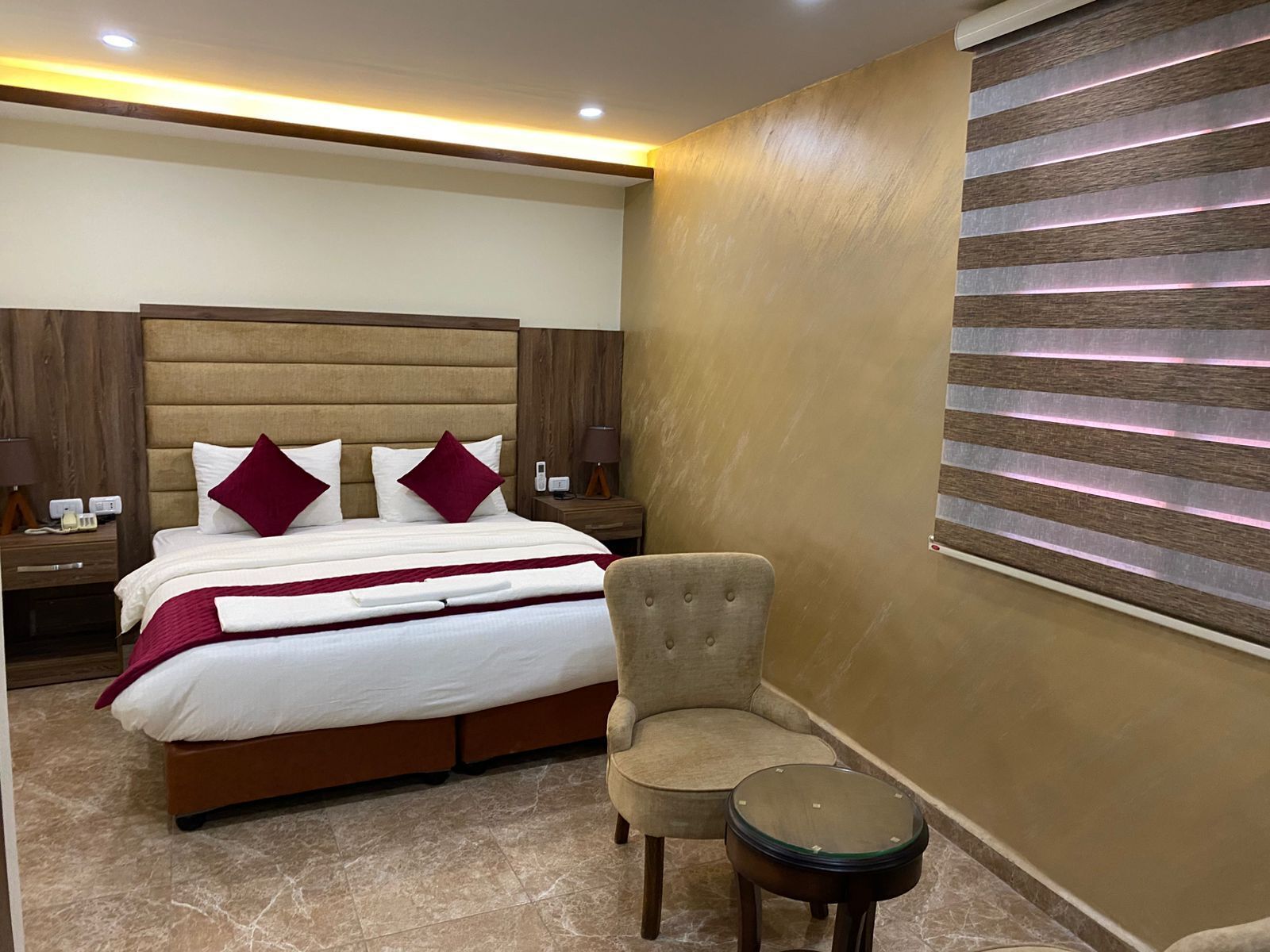 Una camera d'albergo con un letto grande, una sedia, un tavolino e una parete illuminata lateralmente.