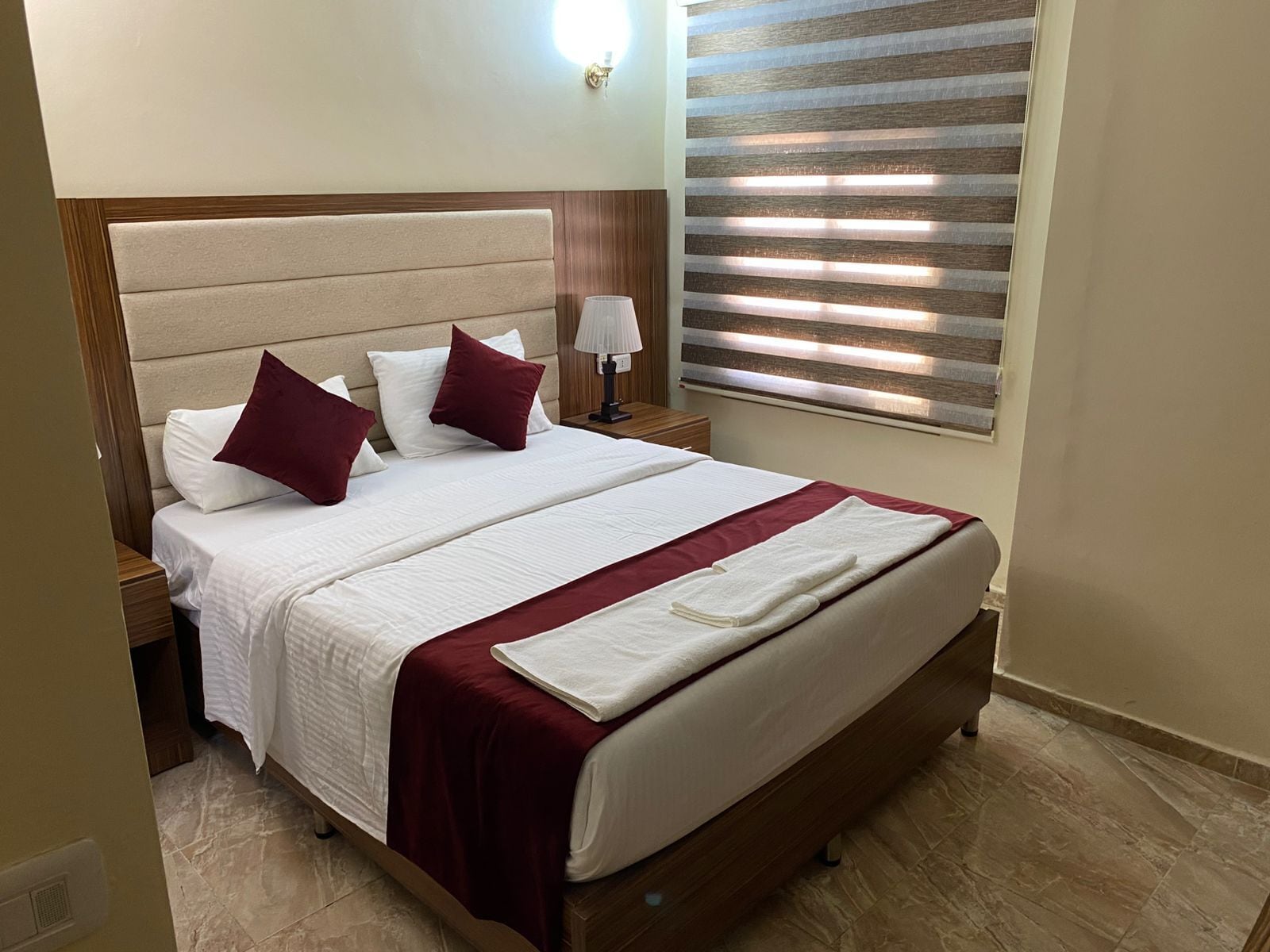 A neatly made bed with white bedding and maroon accents, a bedside lamp, and a window with blinds.