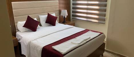 A neatly made bed with white bedding and maroon accents, a bedside lamp, and a window with blinds.