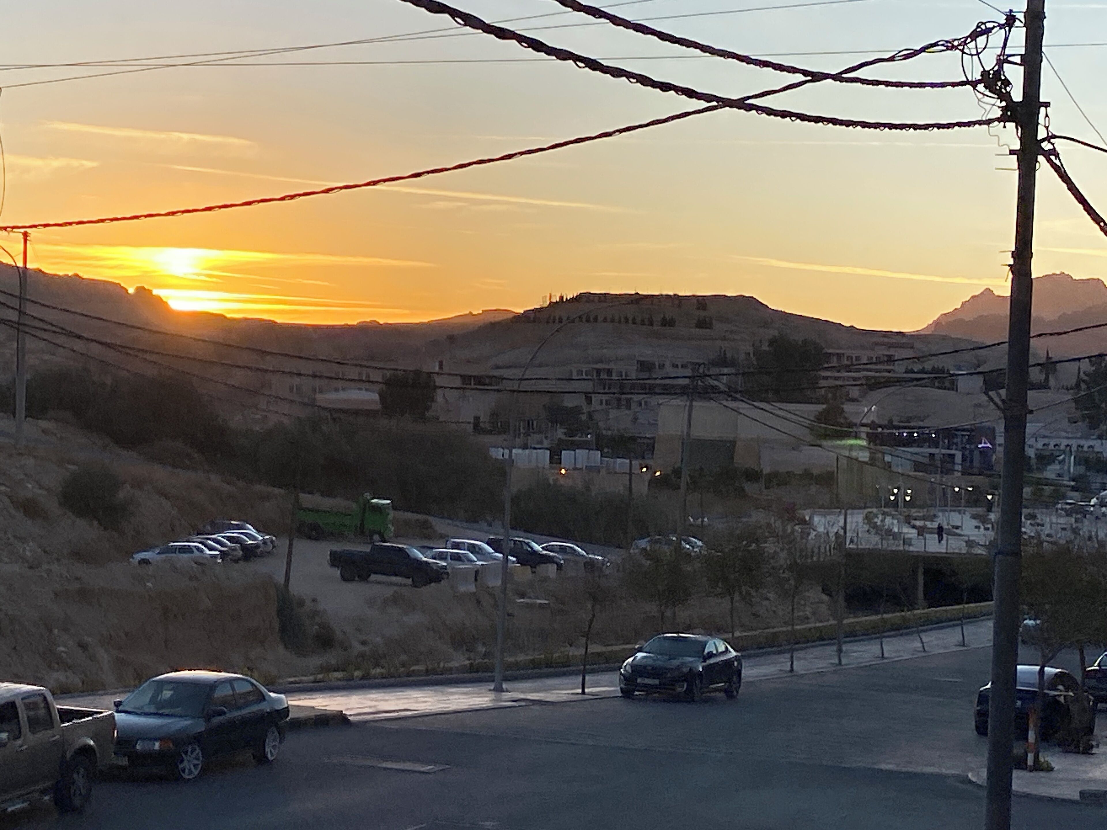 A parking lot with several cars and a few trucks. There are hills in the background and a sunset in the sky.