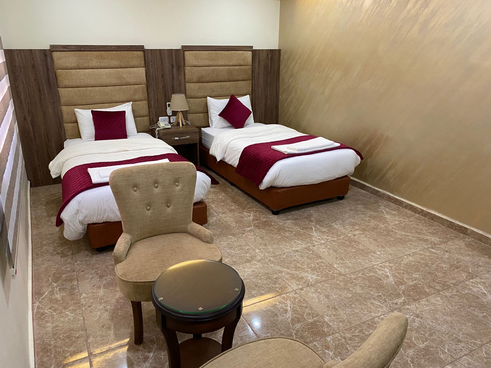 A hotel room with two beds, a chair, a small table, and a nightstand with a lamp.