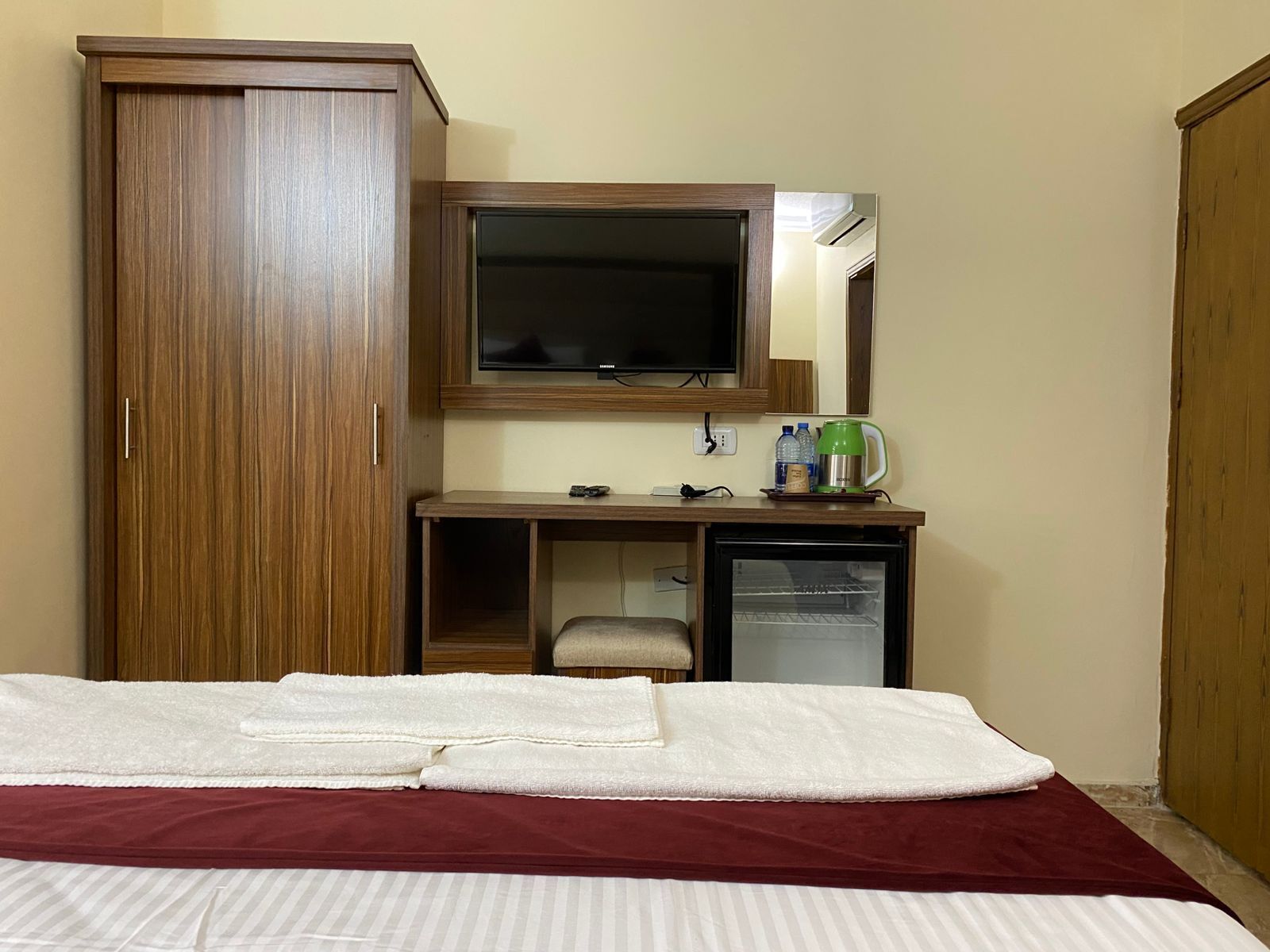 Habitación de hotel con cama, armario, escritorio con espejo, televisor y refrigerador pequeño.
