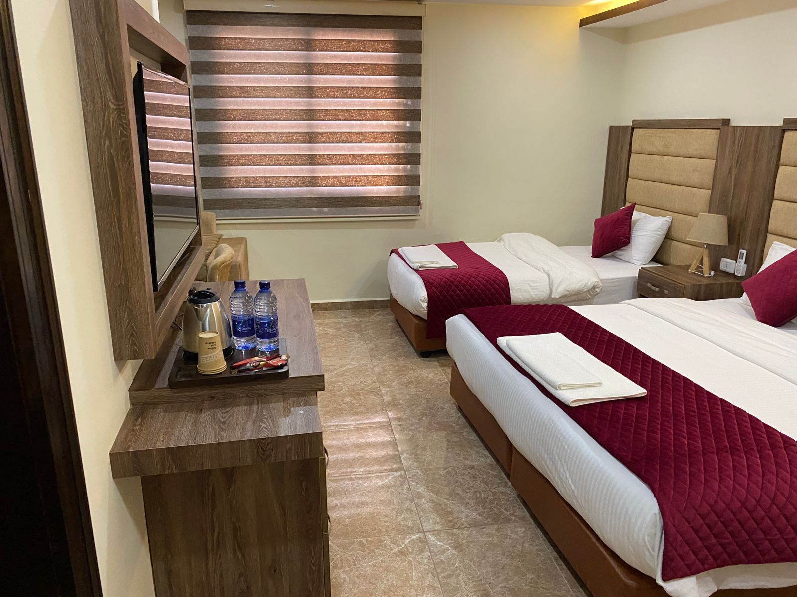 Une chambre d’hôtel avec deux lits, une table de chevet en bois, une petite table avec des bouteilles d’eau et une machine à café, et une fenêtre avec des stores.
