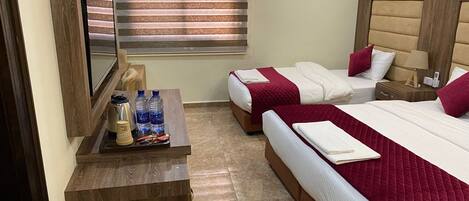 Habitación de hotel con dos camas, una mesita de noche de madera, una pequeña mesa con botellas de agua y una cafetera, y una ventana con persianas.