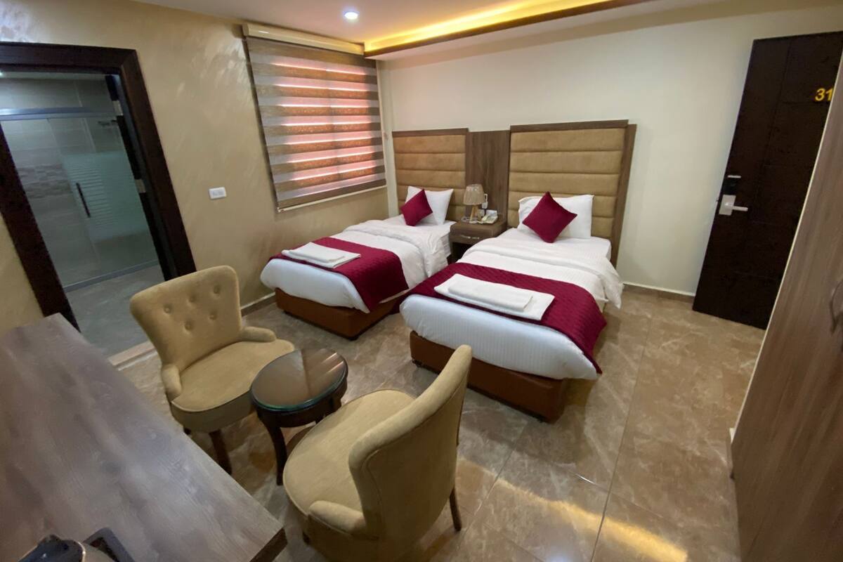 A hotel room with two beds, a small round table, two armchairs, and a glass door leading to a balcony.