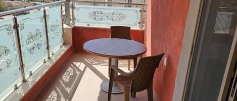 A balcony with a round table and two chairs, a glass railing, and a view of buildings and hills in the background.