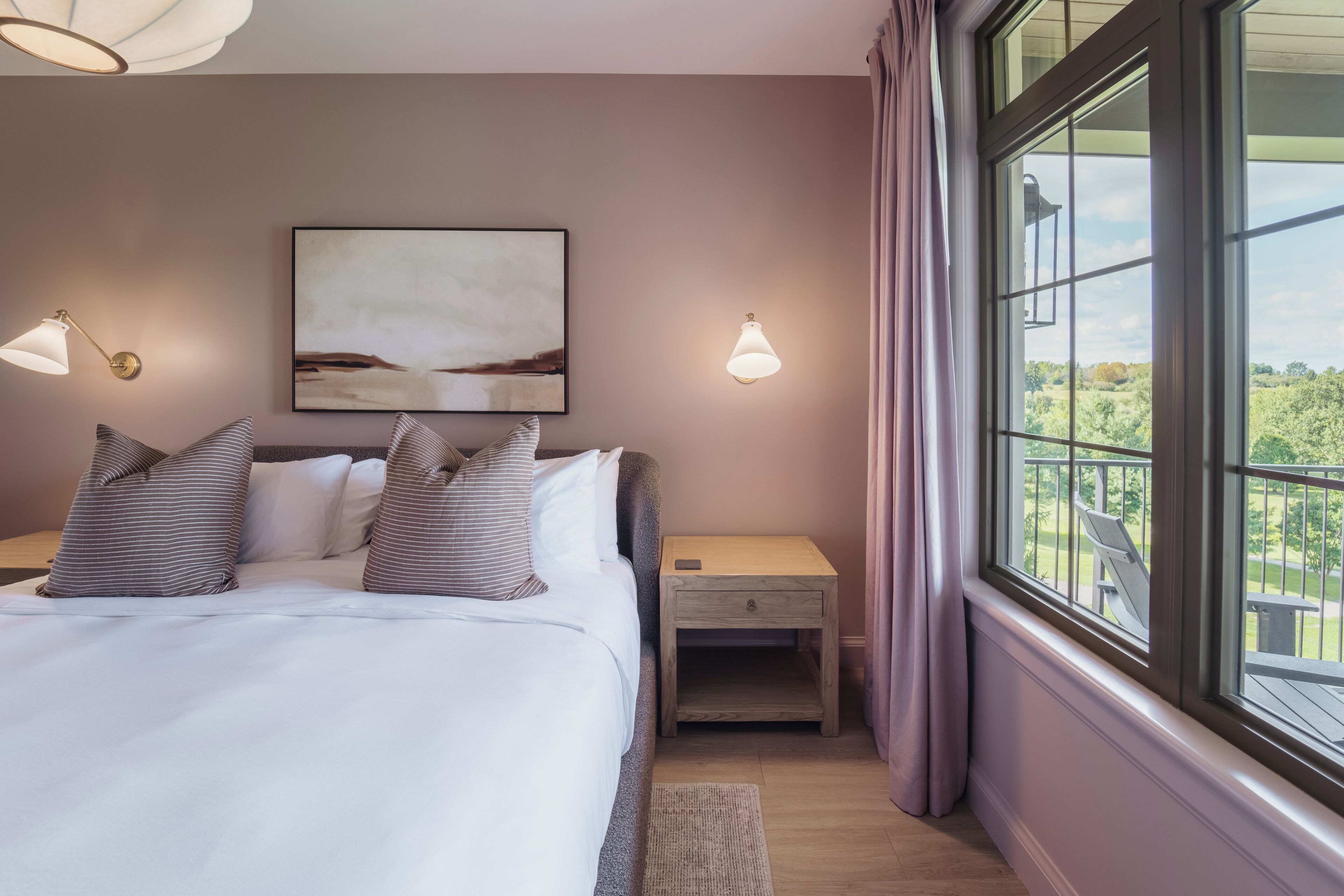 A bedroom with a bed, bedside tables, a nightstand, a painting, and a window with a view.