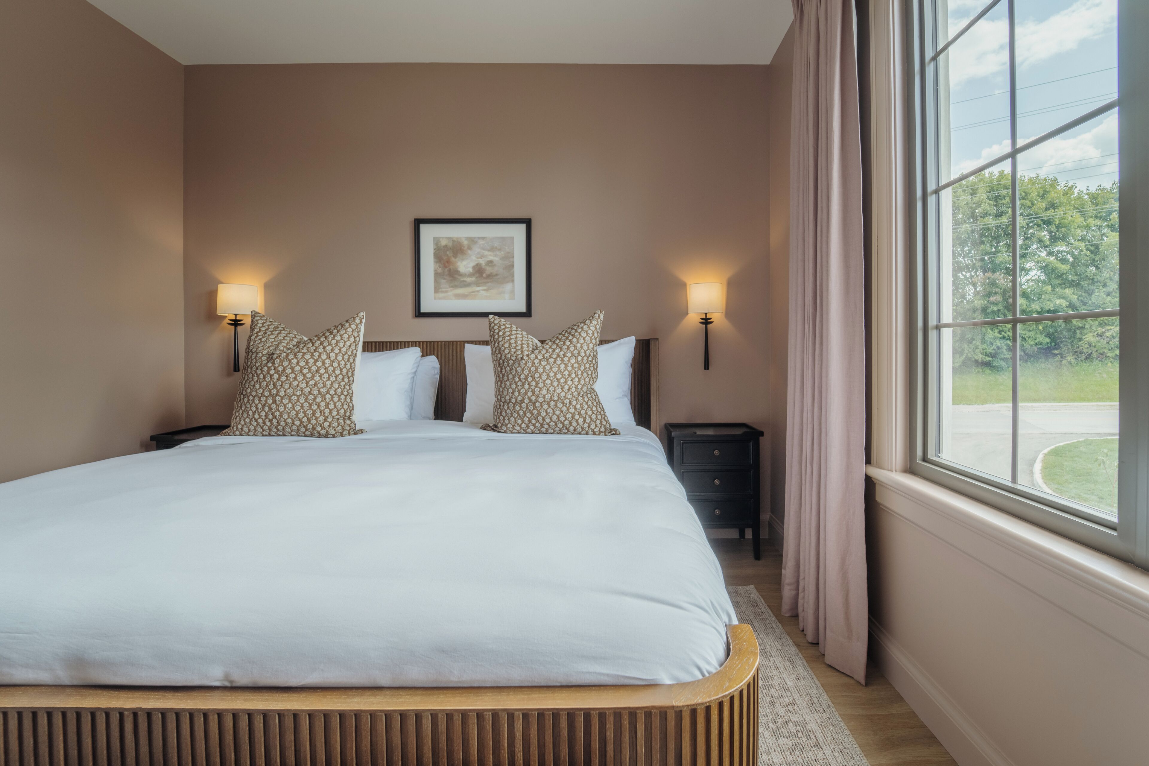 Une chambre à coucher bien rangée, avec un grand lit, deux lampes de chevet, un tableau encadré au mur et une fenêtre avec des rideaux.
