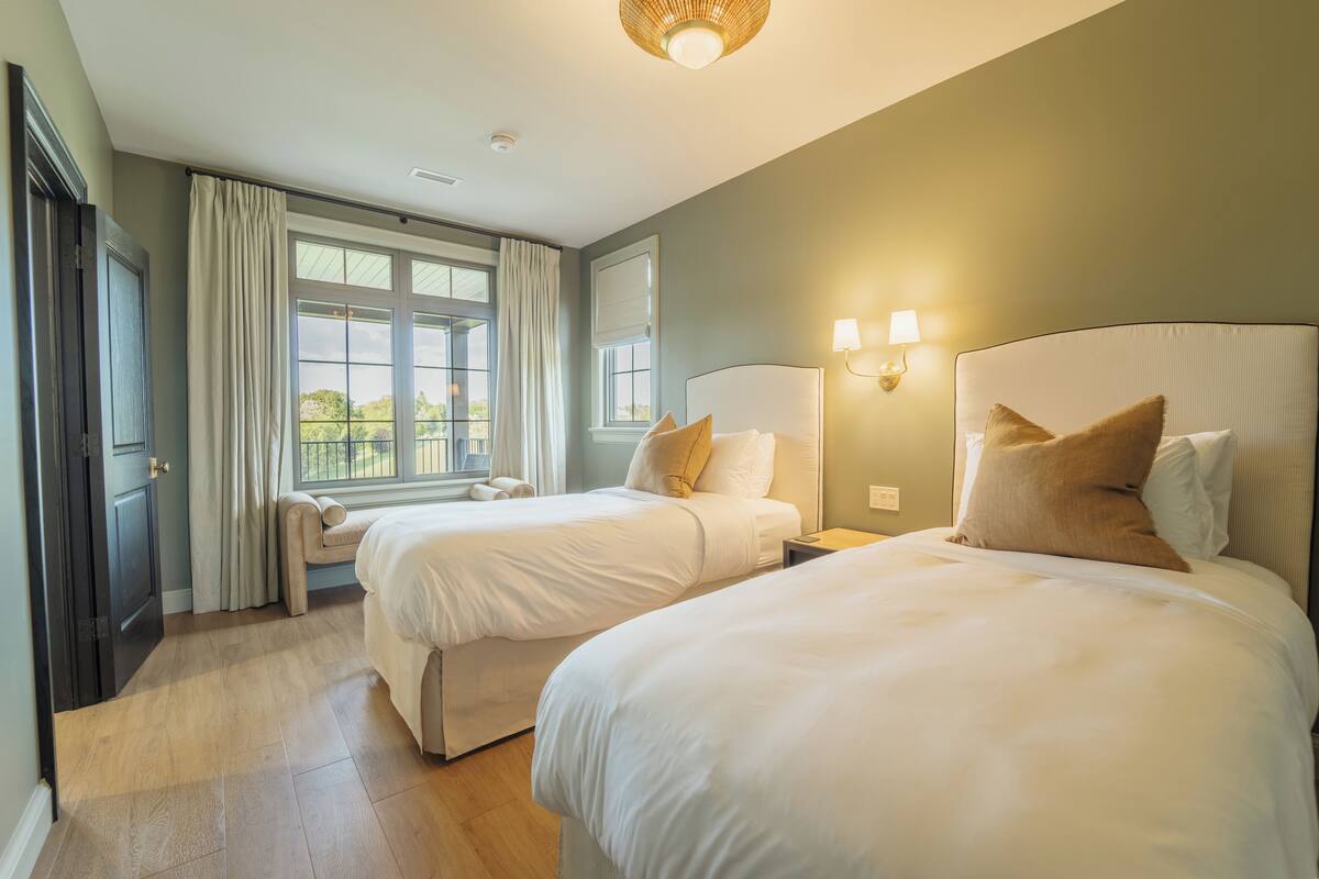 A hotel room with two beds, a window with curtains, a bedside table with a lamp, and a door.