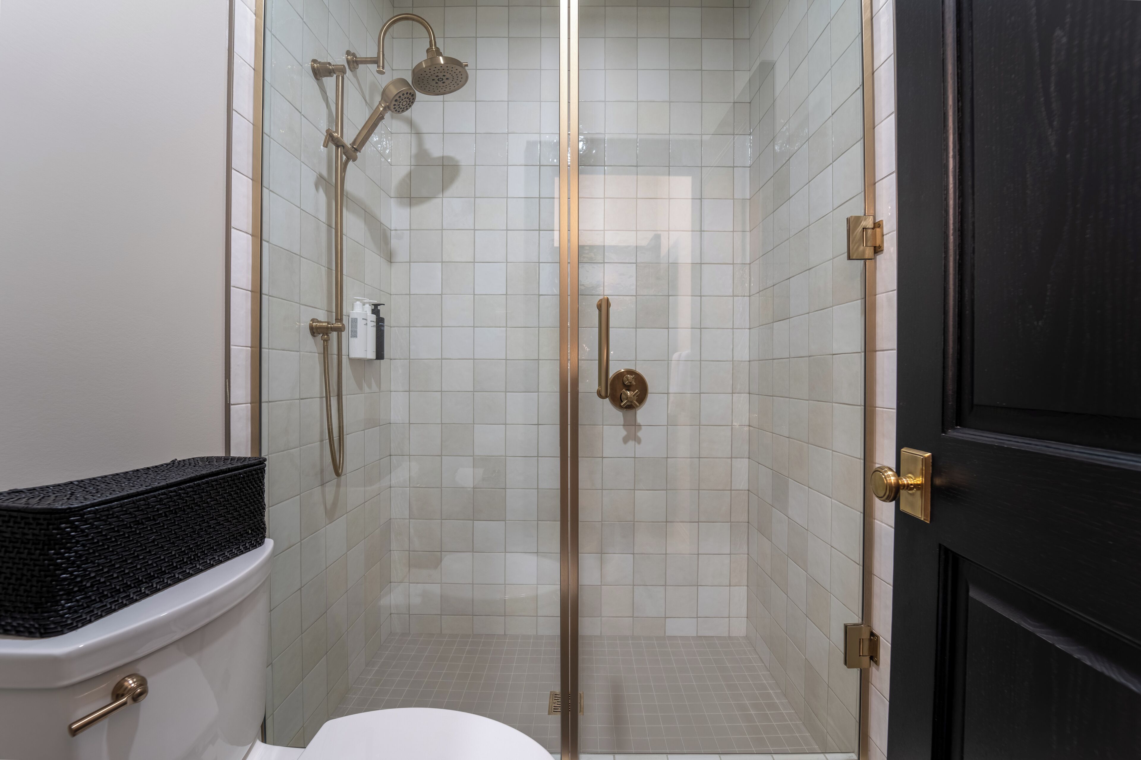 Une salle de bain avec une douche à parois de verre, des toilettes et un panier noir posé sur les toilettes.