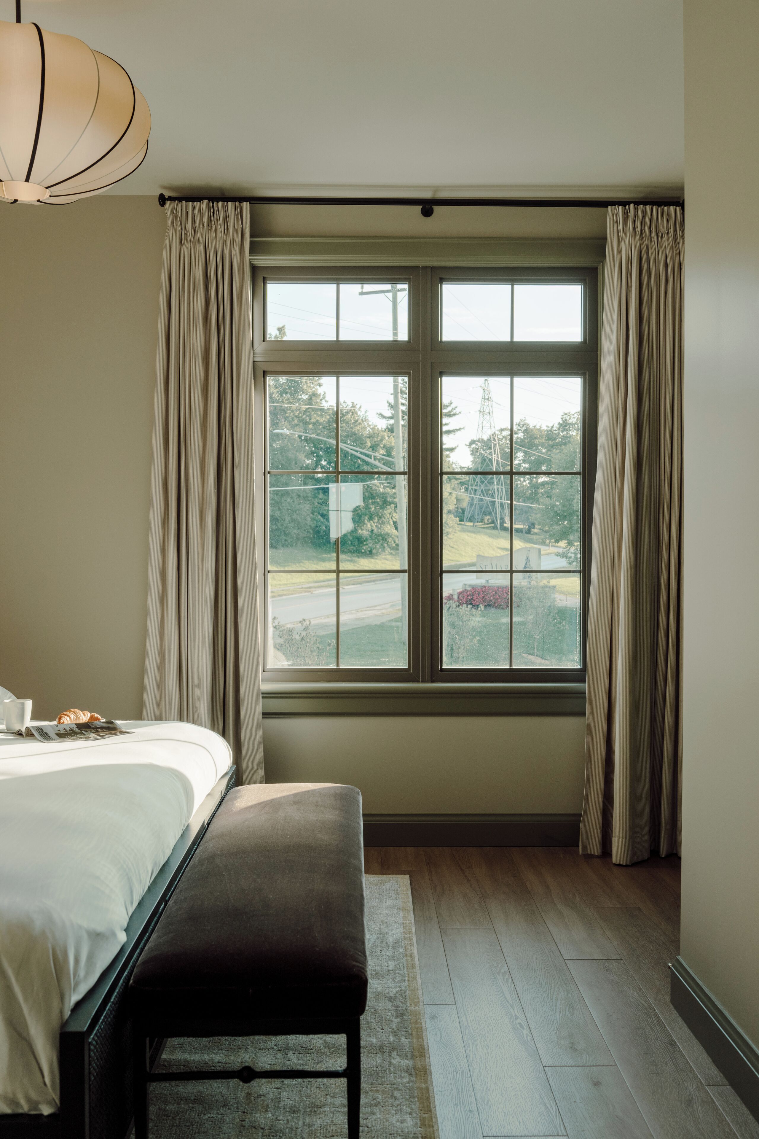 A bedroom with a large window, a bed, a bench, and a hanging light fixture.